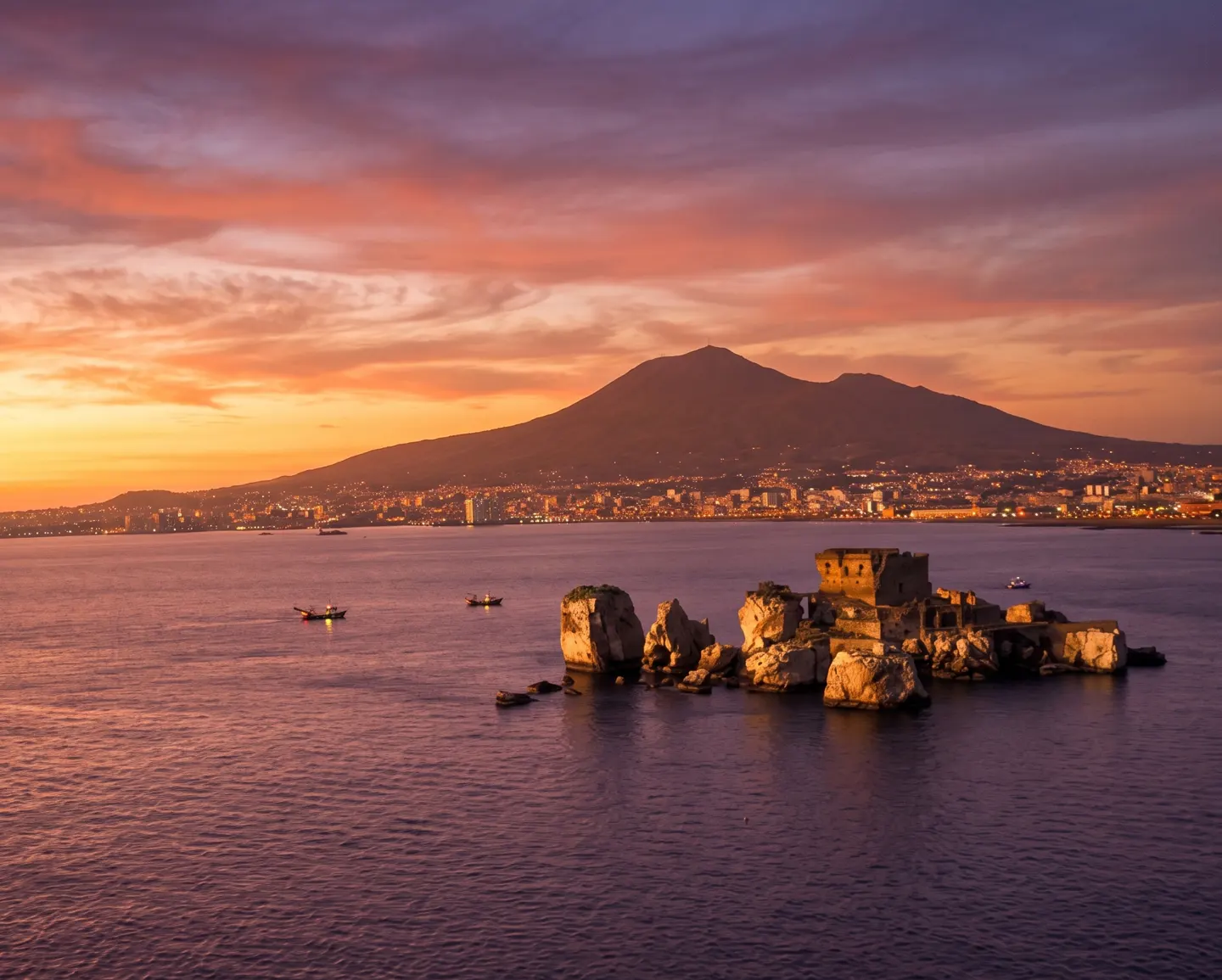A rocky island with ruins sits in the sea at sunset, with Mount Vesuvius and a coastal city in the background.