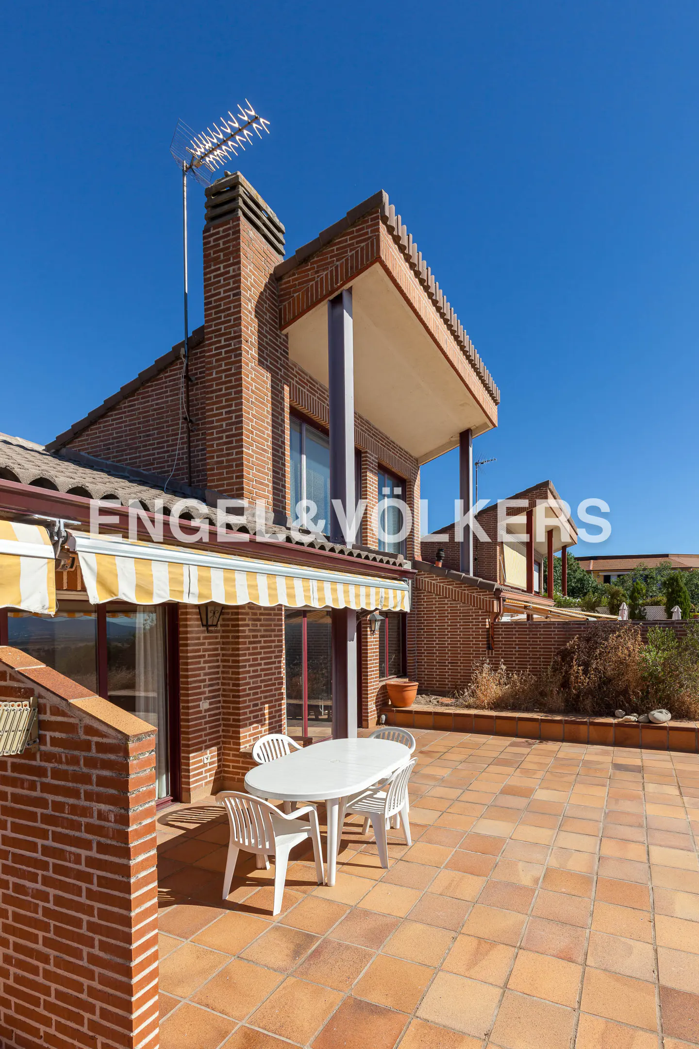 Exterior view of a brick house with a patio, featuring a white table and chairs under a yellow and white striped awning. Blue sky above.