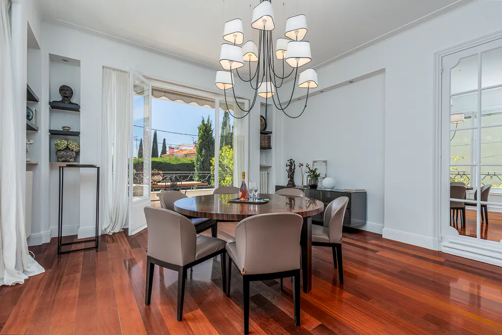 Bright dining room with wood floors, white walls, and a round table with six chairs. A chandelier hangs above the table. Balcony doors open to a view of trees.