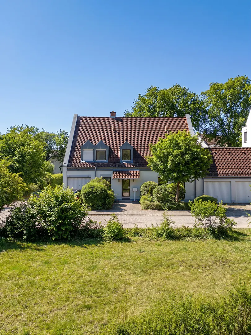 Exterior view of a light blue house with a red tile roof, dormer windows, and green trees against a clear blue sky.