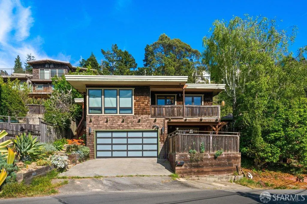 A two-story brown shingle house with a blue garage door and balconies on a sunny day.