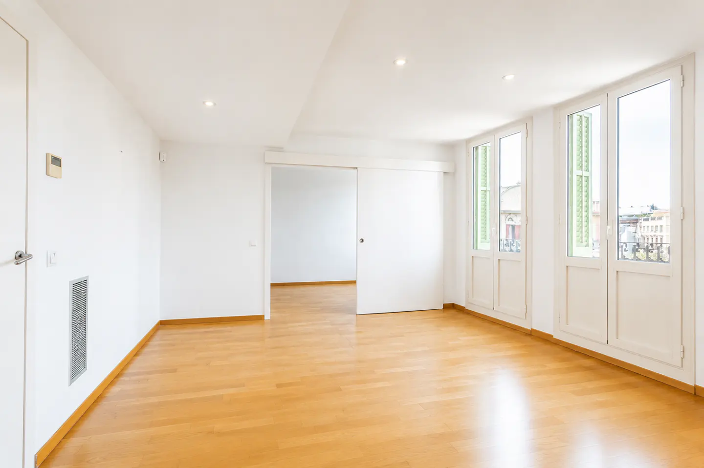 Bright, empty room with light wood floors, white walls, a sliding door, and large windows with green shutters.