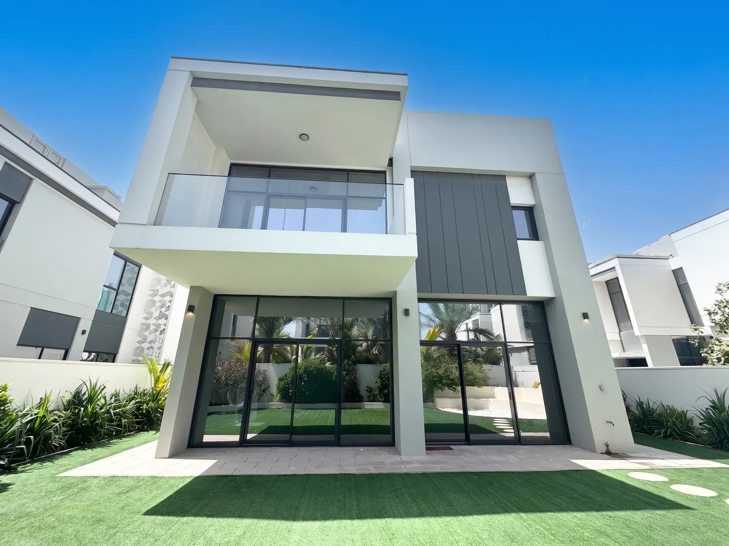 Modern two-story house with a balcony, large windows, and a gray and white exterior, set against a clear blue sky.