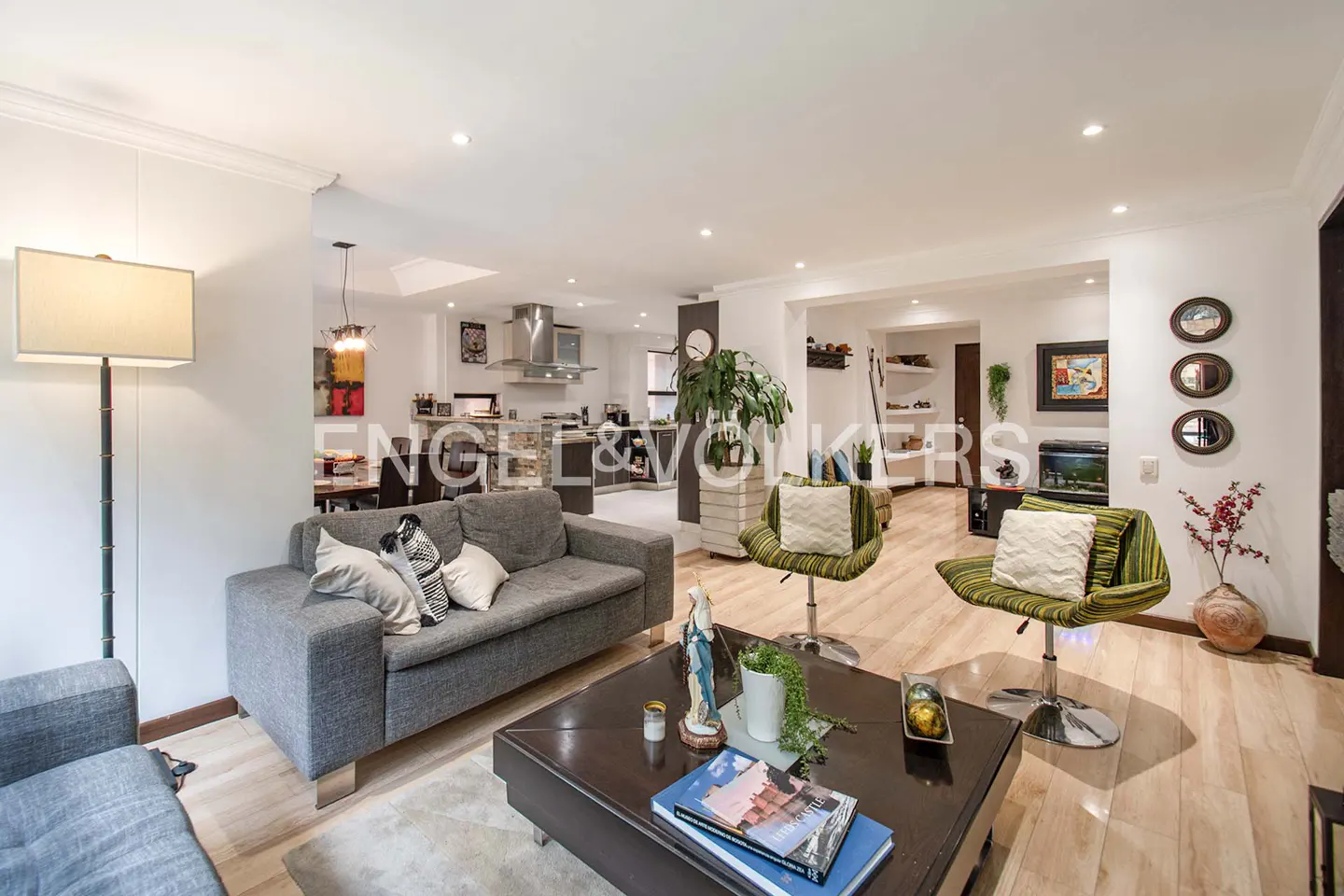 Open-concept living room with gray sofas, striped chairs, and a dark wood coffee table on light wood floors. Kitchen in background.