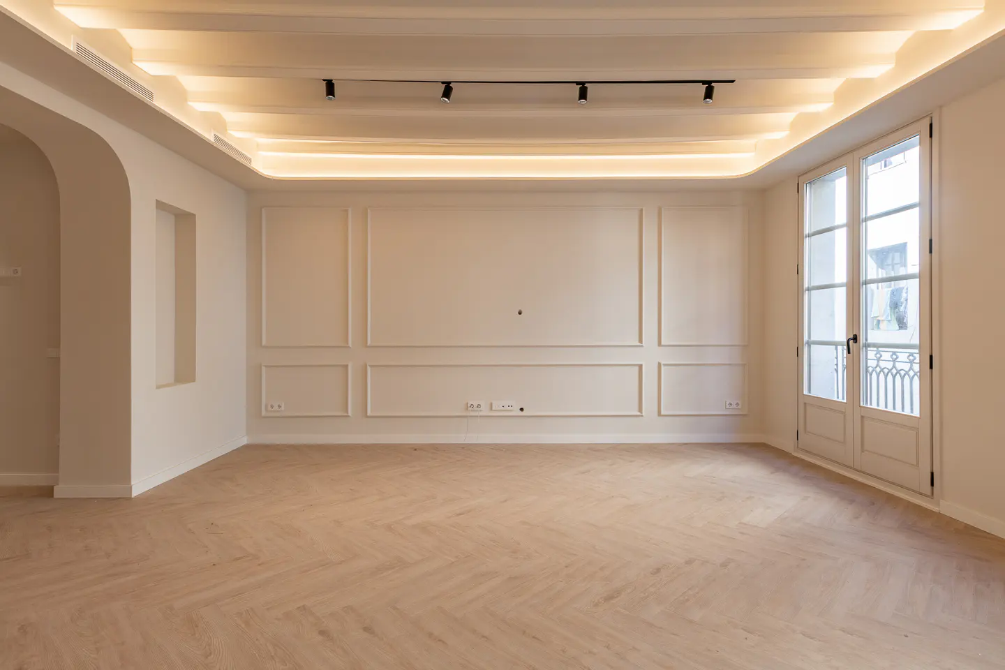 Bright, empty room with herringbone wood floors, white walls with molding, and a white ceiling with recessed lighting. Double doors to the right.