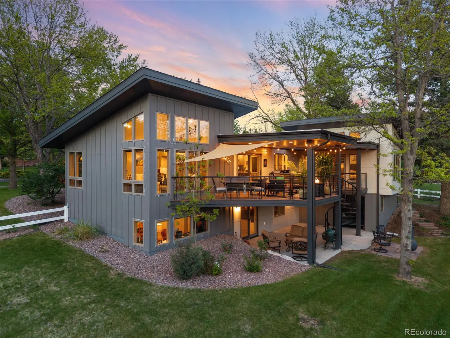 Two-story gray house with a deck, outdoor furniture, and a spiral staircase. Green lawn and trees surround the house. Sunset sky.