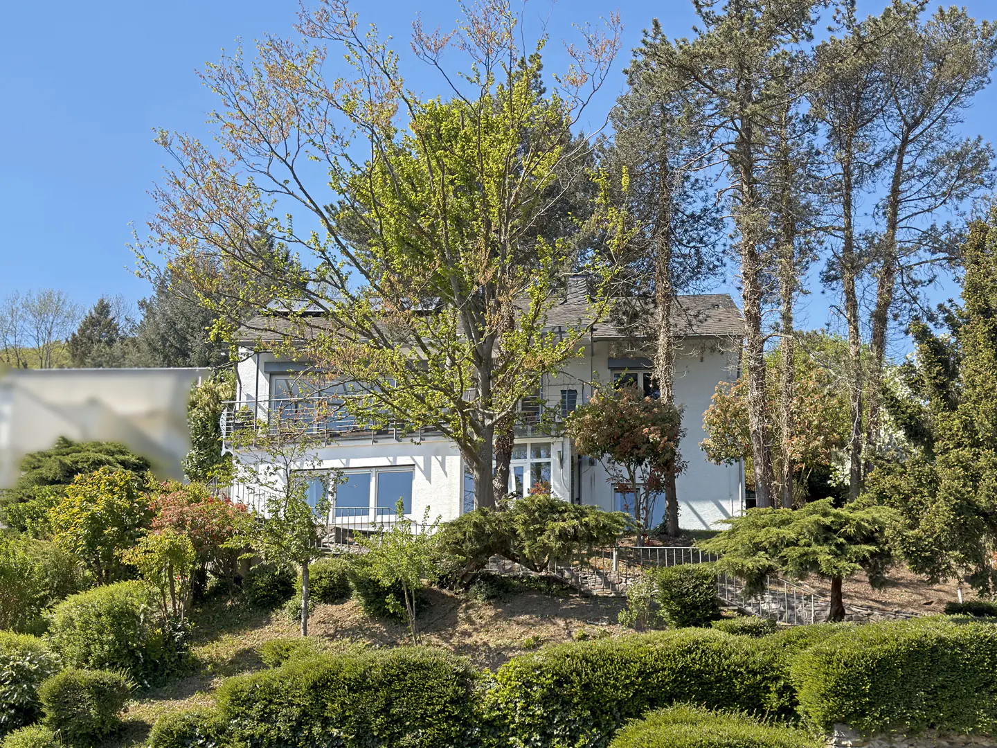 Two-story white house with a balcony, surrounded by trees and green bushes under a blue sky.