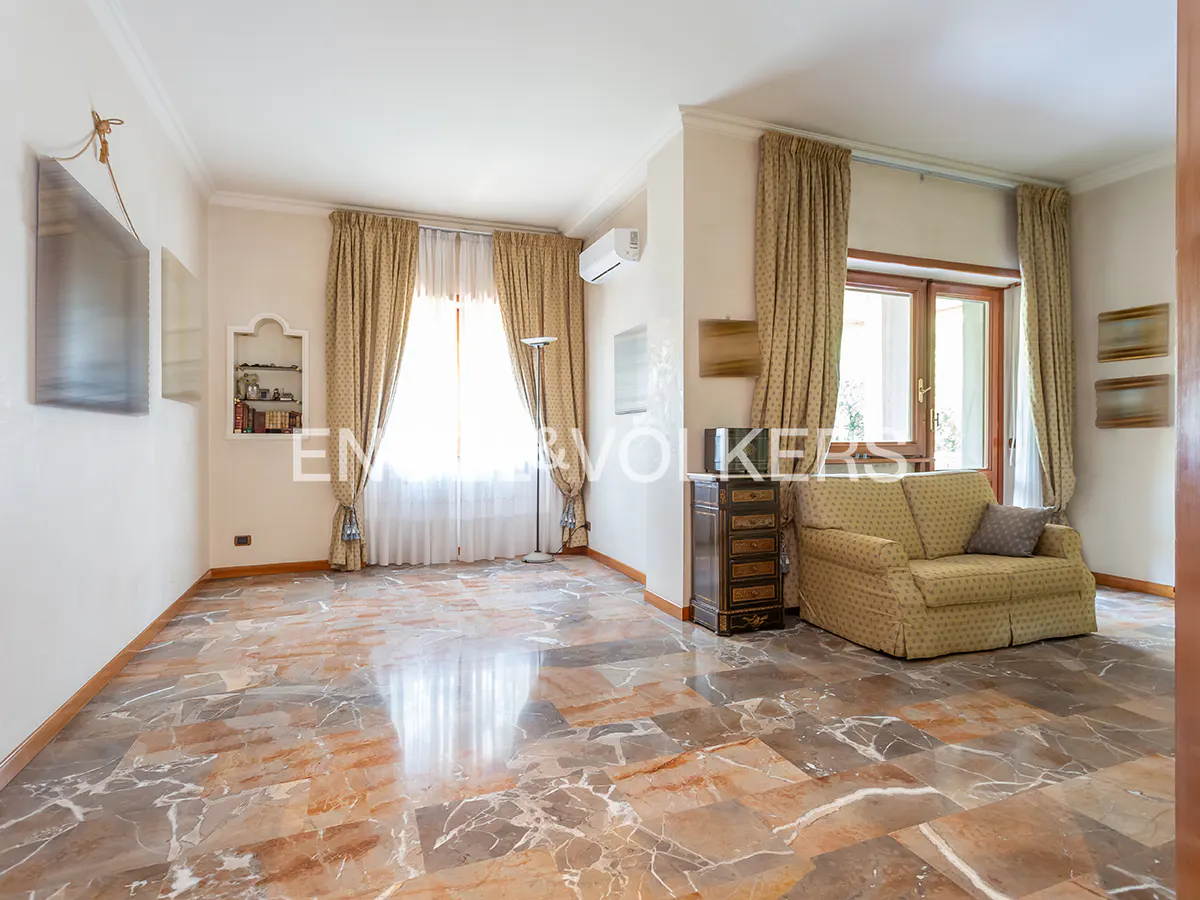A living room with marble floors, beige walls, and patterned curtains. A tan sofa sits near a dark wood cabinet. Natural light streams through the windows.
