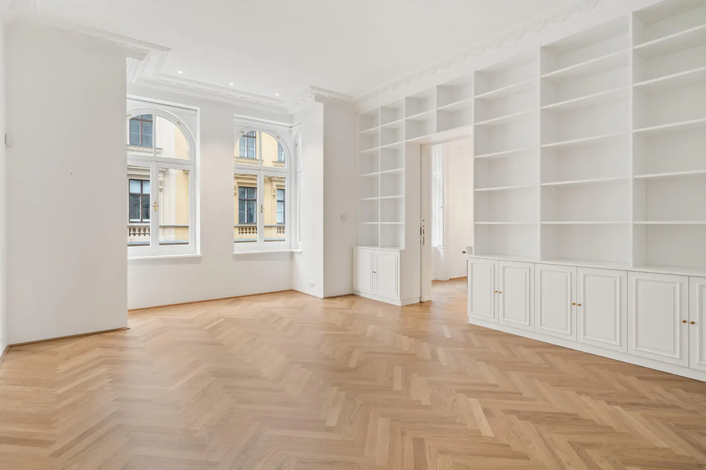 Bright, empty room with herringbone wood floors, white walls, arched windows, and built-in white bookshelves and cabinets.