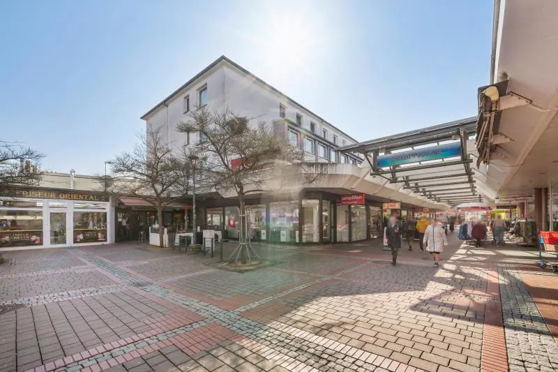 Outdoor view of a shopping center with stores, people walking, and a brick-paved pedestrian area on a sunny day.