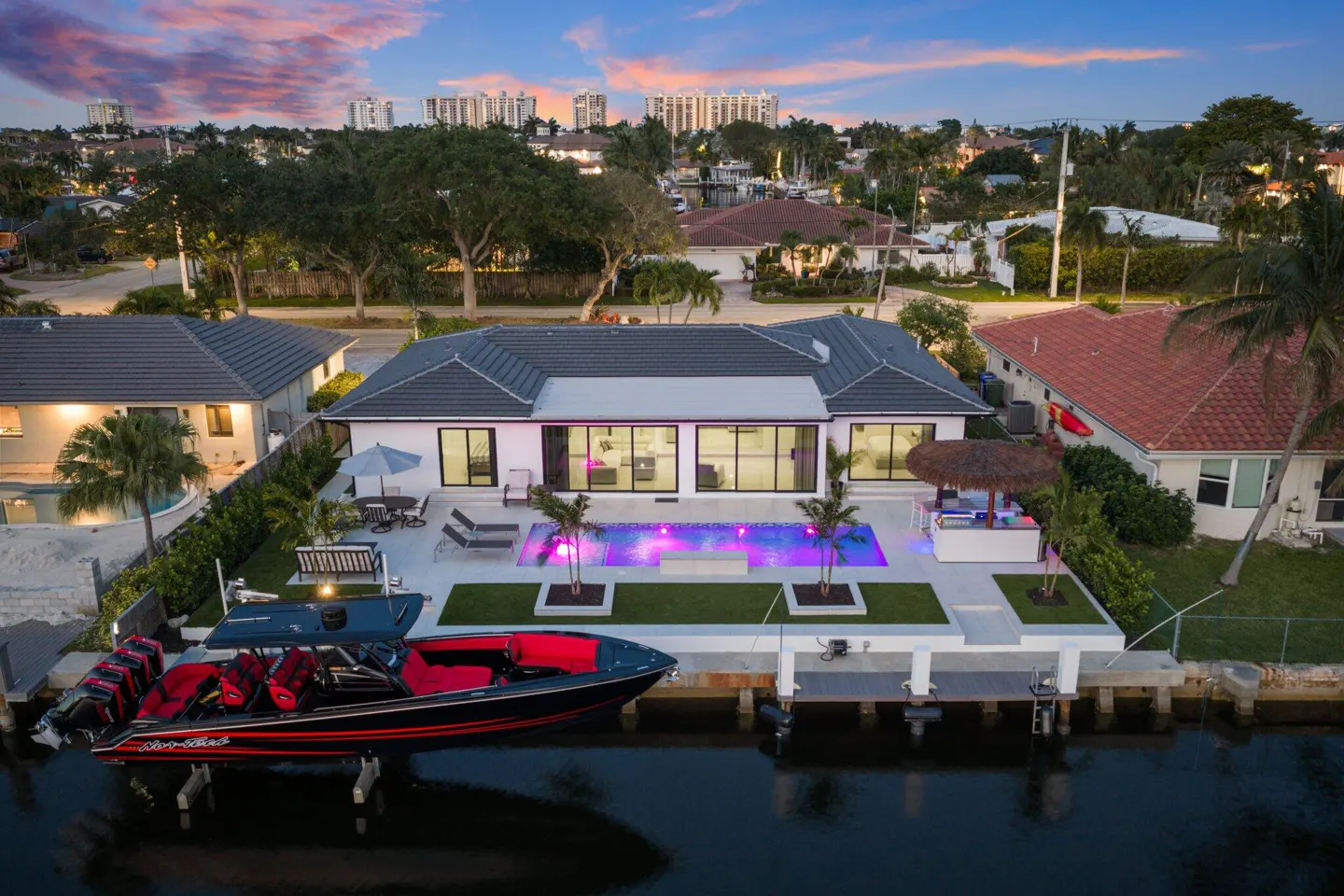 Aerial view of a modern white waterfront home with a pool, tiki bar, and a black and red speedboat docked.