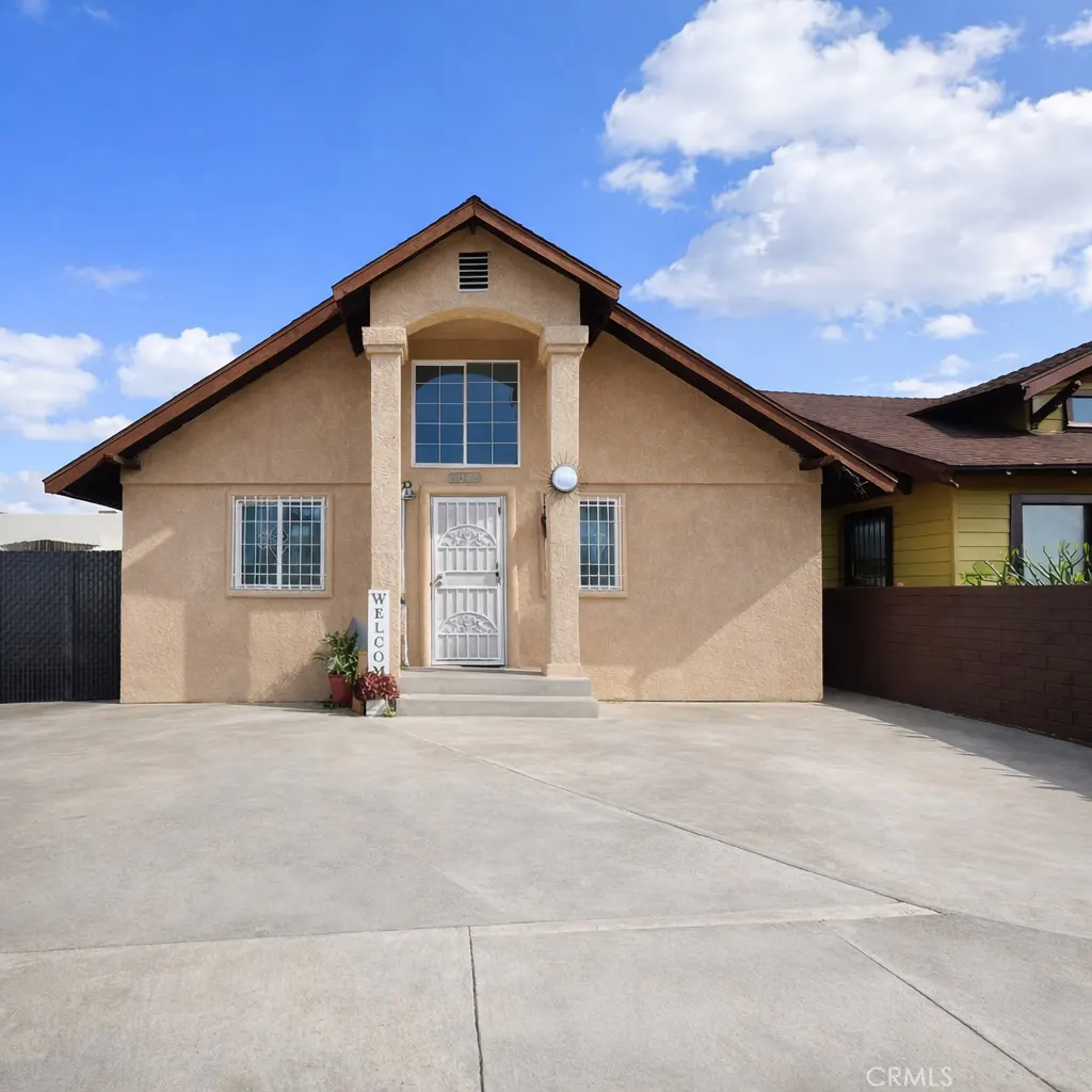 Front view of a one-story beige house with a white door, columns, and a "Welcome" sign on the porch. Blue sky with clouds above.
