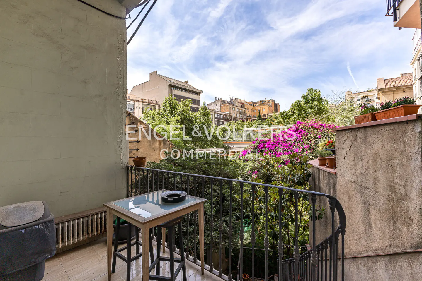 Balcony view with table, chairs, and black railing overlooking lush greenery and city buildings under a cloudy sky.