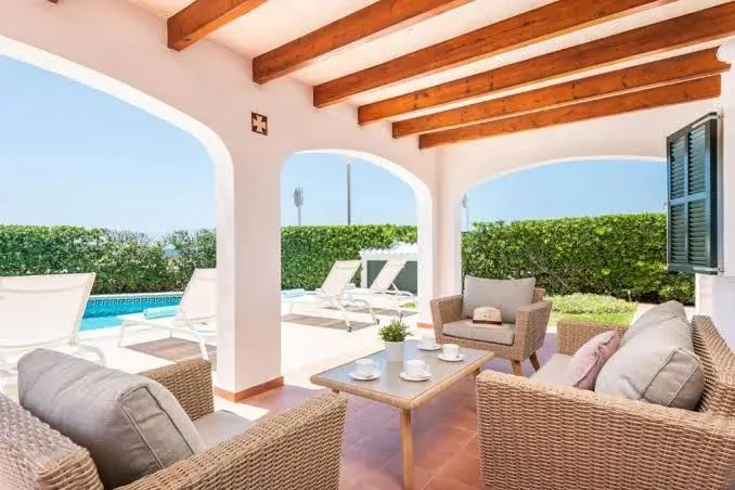 Covered patio with wicker furniture, a coffee table with cups, and a view of a pool and green hedges.