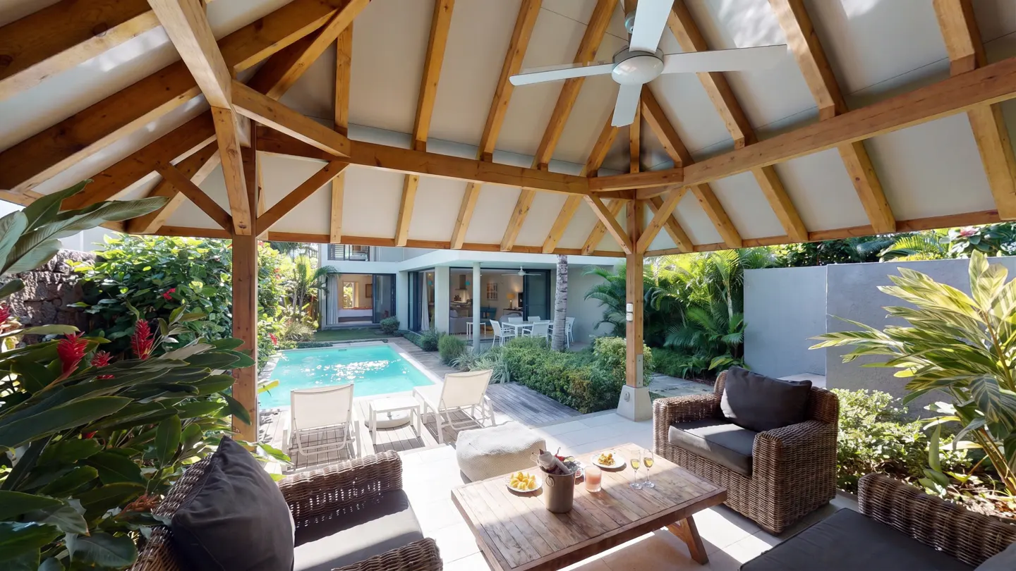 Outdoor patio with wicker furniture under a wooden pergola with a ceiling fan. A pool and lounge chairs are visible in the background.
