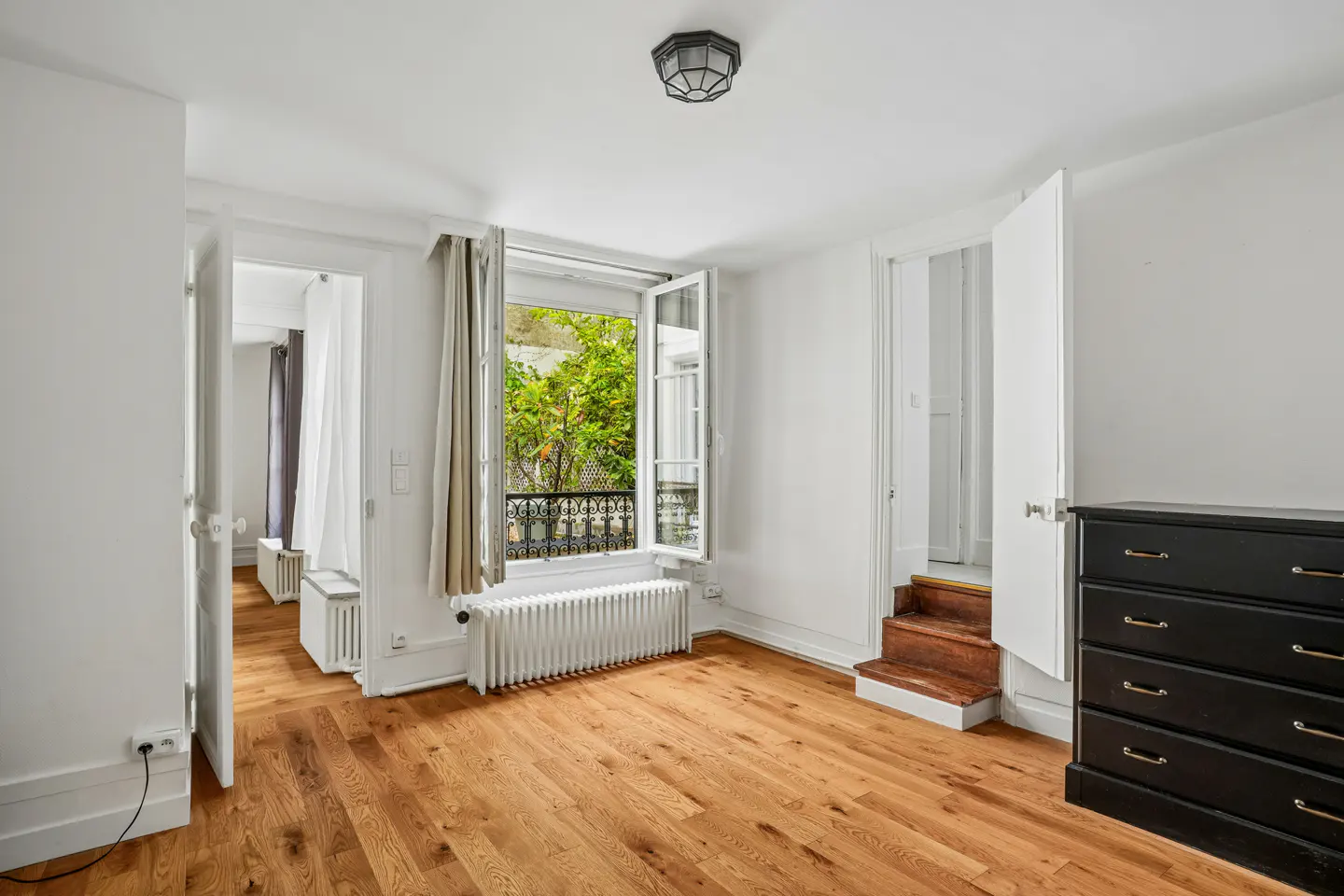 Bright room with hardwood floors, white walls, and an open window showing green trees. A black dresser sits to the right.