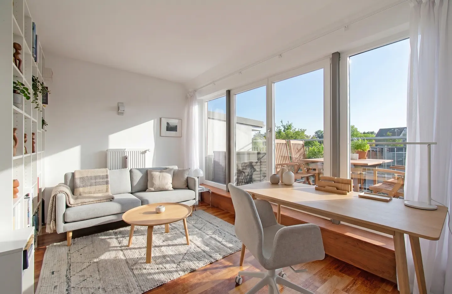 Bright living room with a gray sofa, wooden table, and large windows leading to a balcony with outdoor furniture.