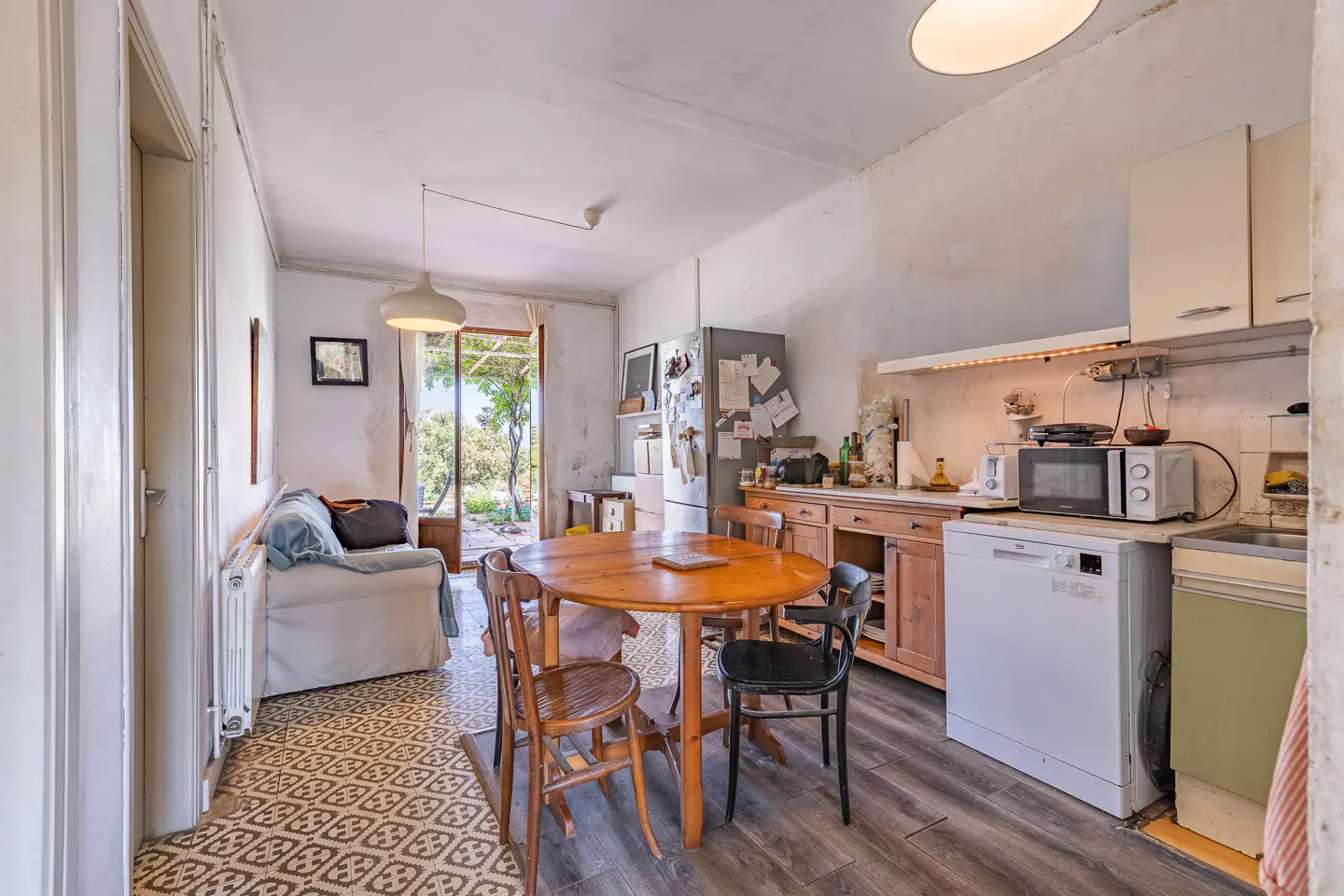 A bright kitchen with a round wooden table and chairs, a white sofa, and an open door to a garden.