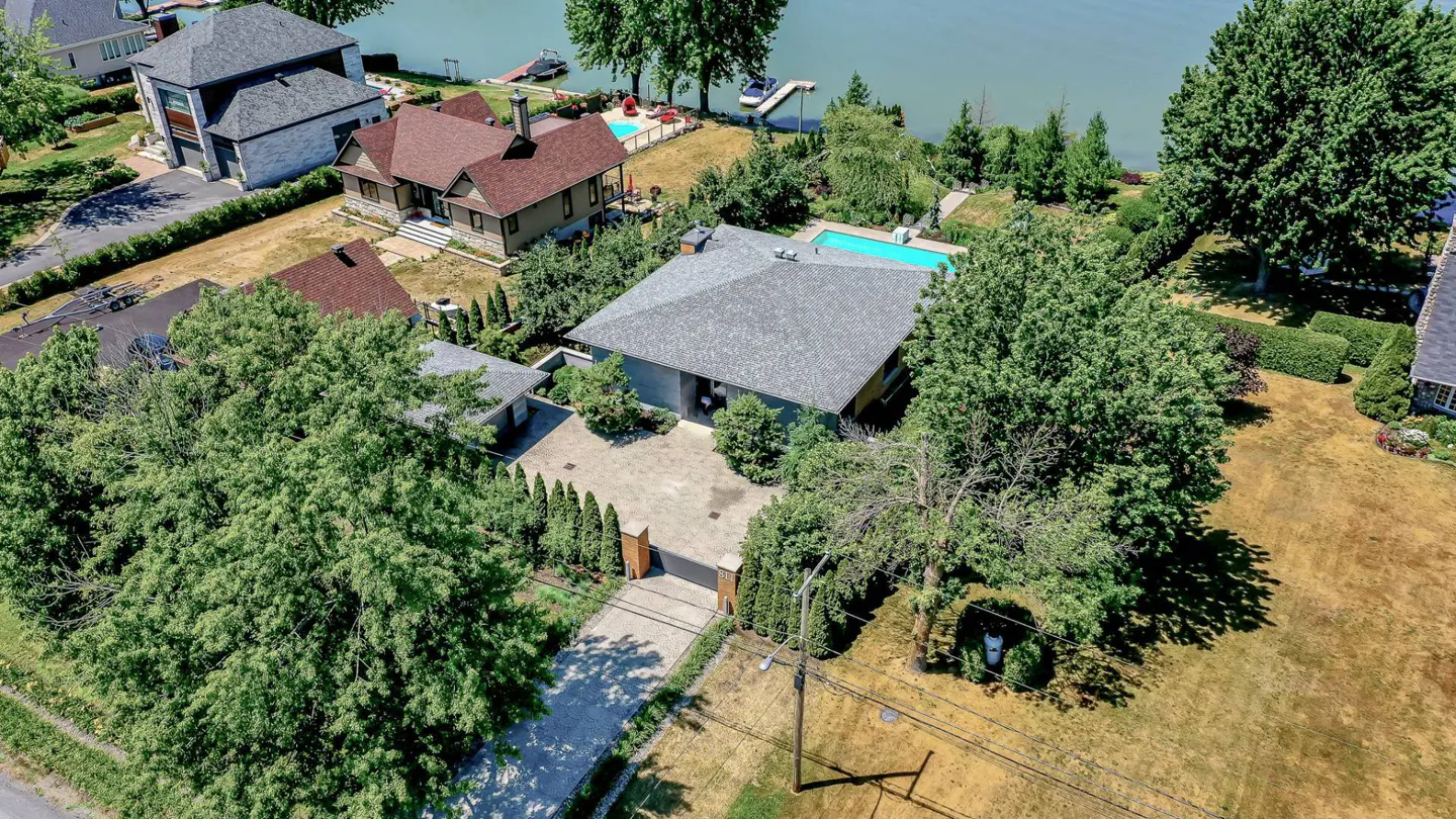 Aerial view of a modern gray house with a gray roof, a stone driveway, and green trees. In the background is a lake and other houses.
