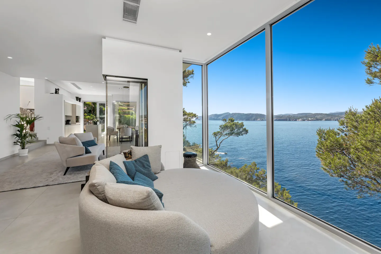 Bright, modern living room with a round sofa and ocean view through large windows. Blue pillows accent the neutral tones.