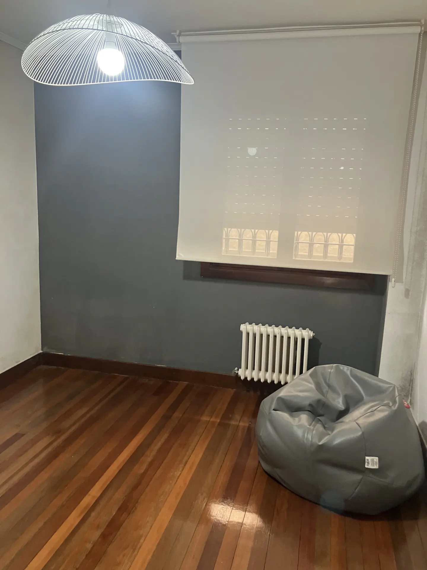 Empty room with wood floors, gray walls, white radiator, and gray beanbag chair. A white shade covers the window.