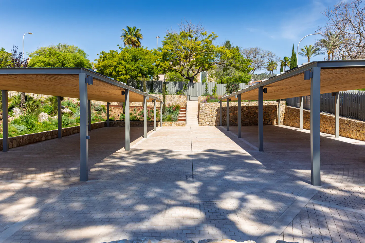 Outdoor parking area with gray metal canopies and straw roofing, stone walls, and a staircase leading to a garden.