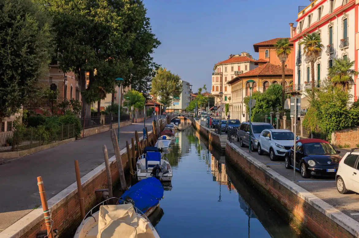 A canal in Treviso, Italy, lined with boats and buildings under a blue sky. Cars are parked along the street.