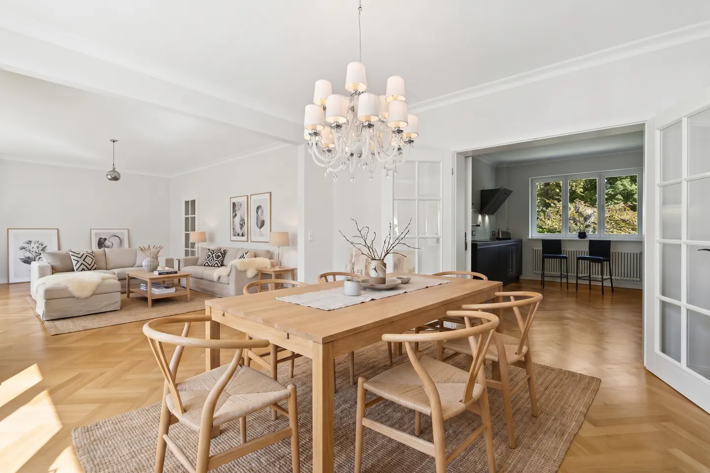 Bright, open-concept living space with a wooden dining table, chairs, and a chandelier. A living room and kitchen are visible in the background.