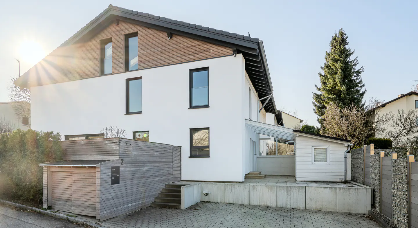 Exterior view of a modern two-story house with white walls, wood accents, and black-framed windows under a sunny sky.