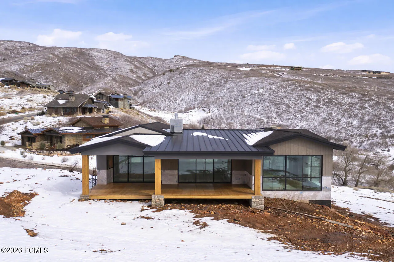 Exterior view of a modern home with a black roof, large windows, and a wooden porch, set against a snowy mountain backdrop.