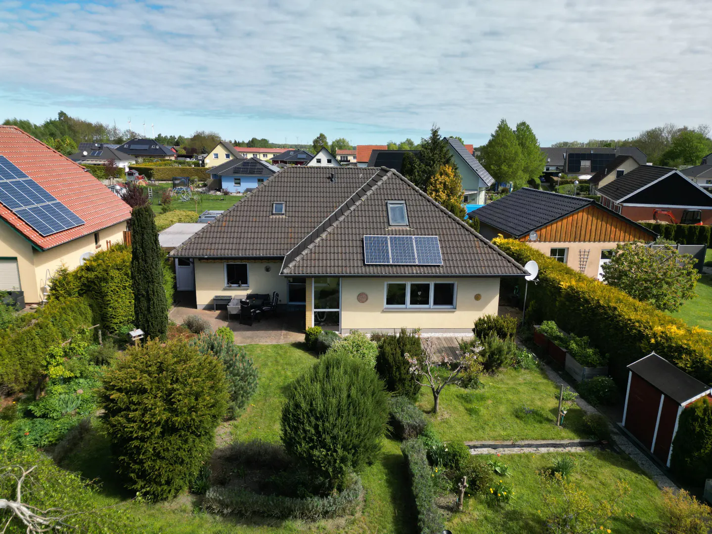 Aerial view of a beige single-story house with a brown roof and solar panels, surrounded by a green lawn and various plants.