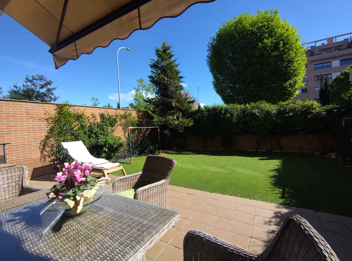 Patio with wicker table, chairs, and pink flowers. A lawn, trees, and a lounge chair are in the background.