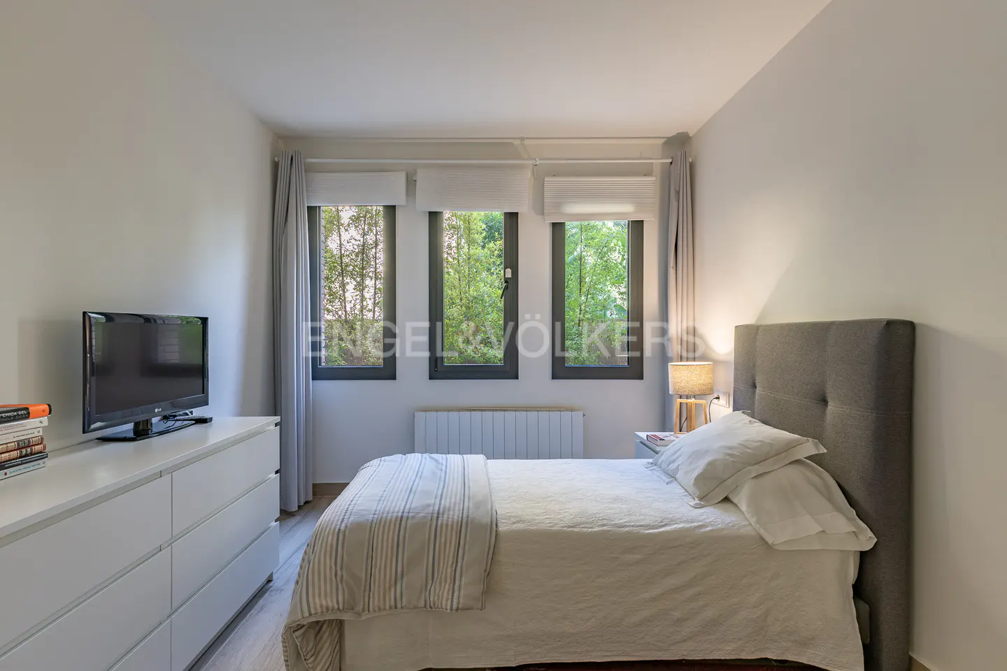 Bedroom with a gray headboard bed, white bedding, three windows with green trees outside, and a white dresser with a TV.