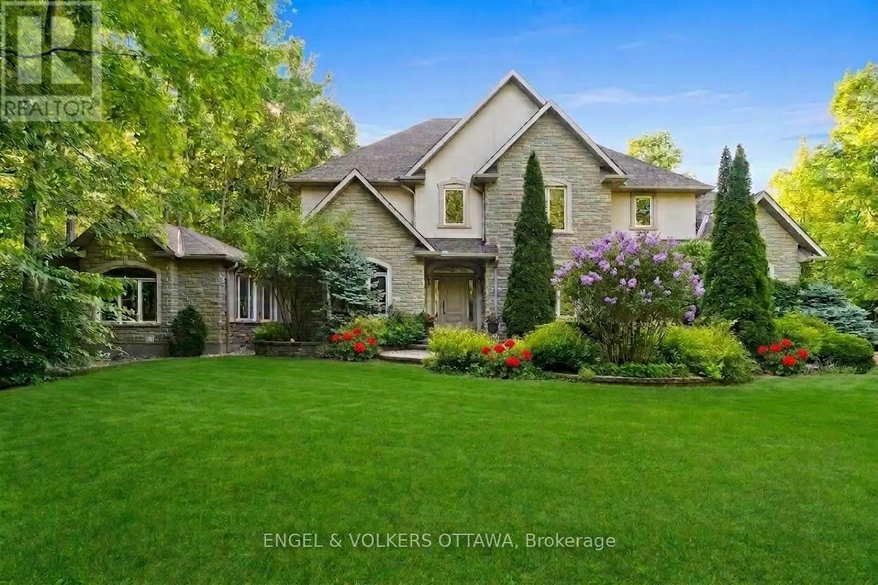 Large stone house with a green lawn, blooming purple lilacs, and red flowers, surrounded by lush trees under a blue sky.