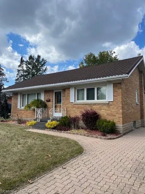 A single-story brick house with a brown roof, white trim, and a brick driveway. Landscaped yard with green grass and colorful bushes. Cloudy sky.