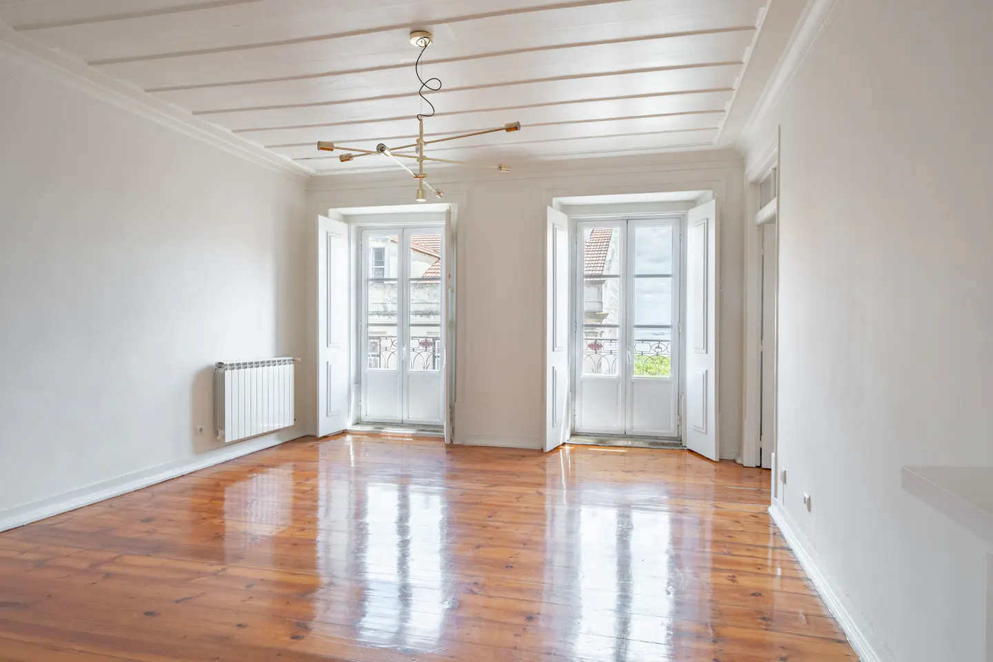 Bright, empty room with wood floors, white walls, and two sets of open French doors. A modern chandelier hangs from the white, beamed ceiling.