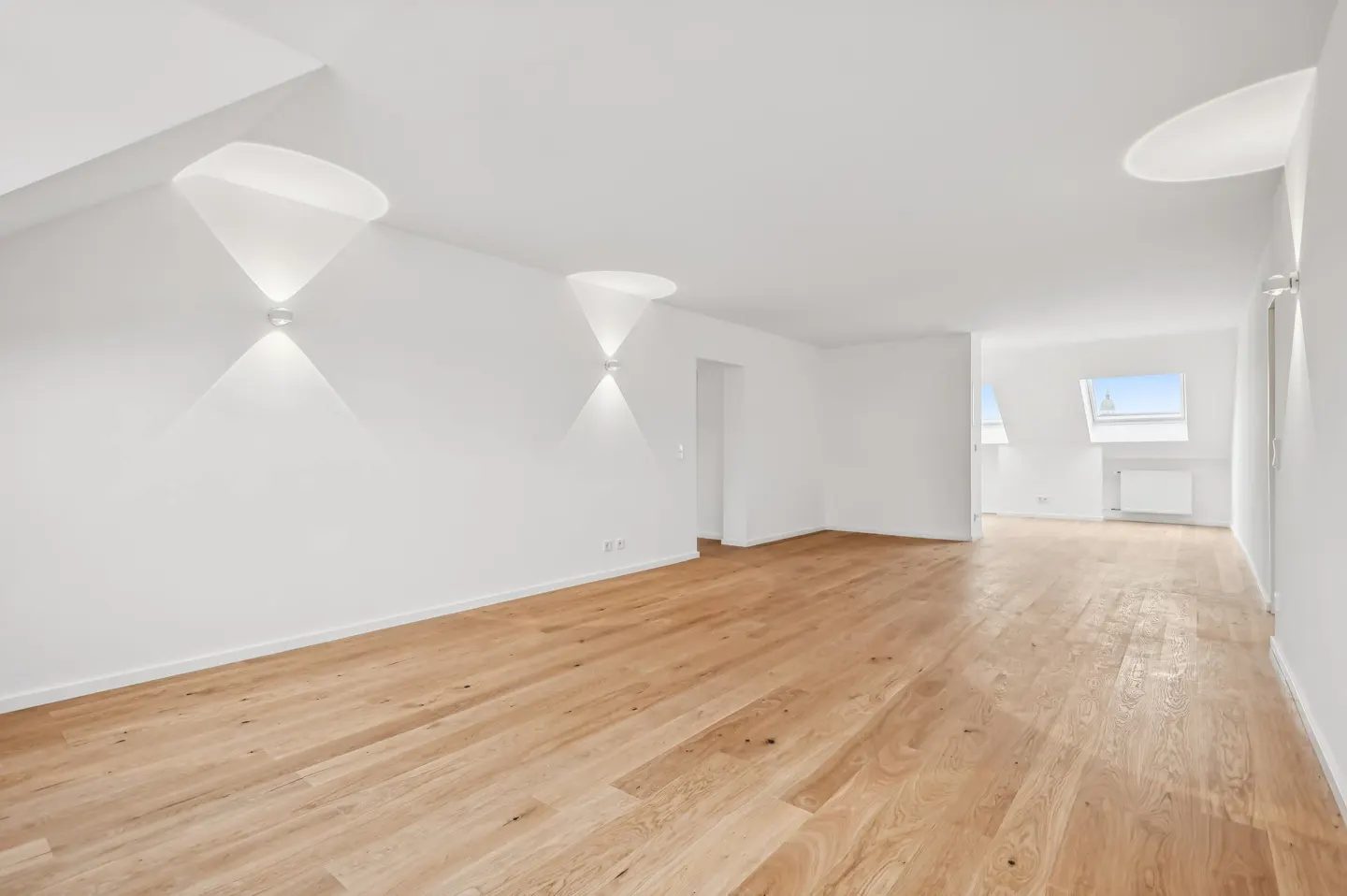 Bright, empty room with light wood floors and white walls. Modern sconces and skylights add architectural detail.