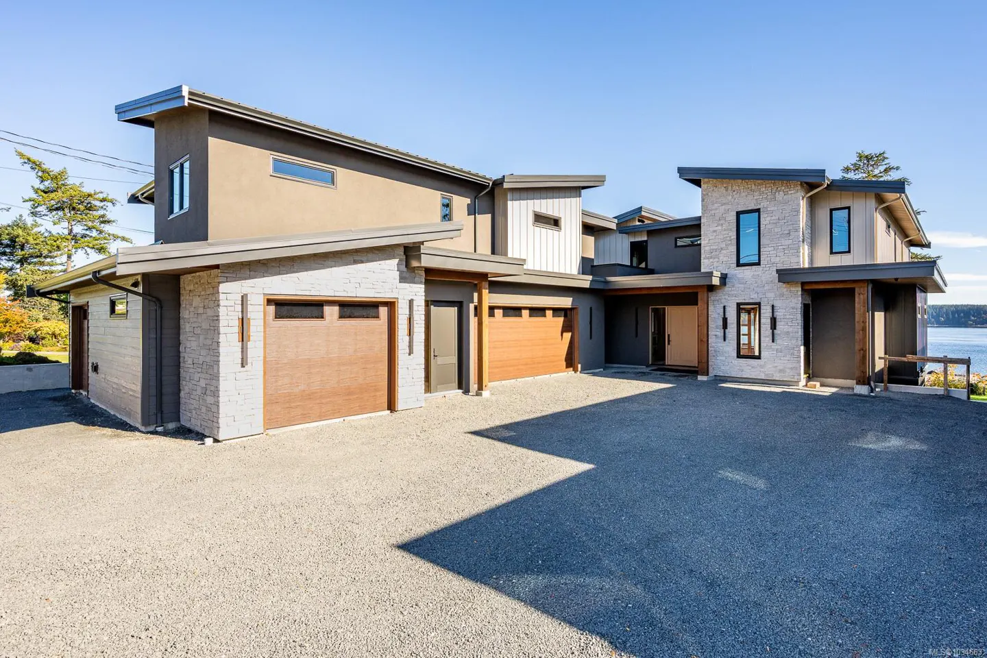 Modern home exterior with stone and stucco, featuring wood garage doors and a gravel driveway on a sunny day.