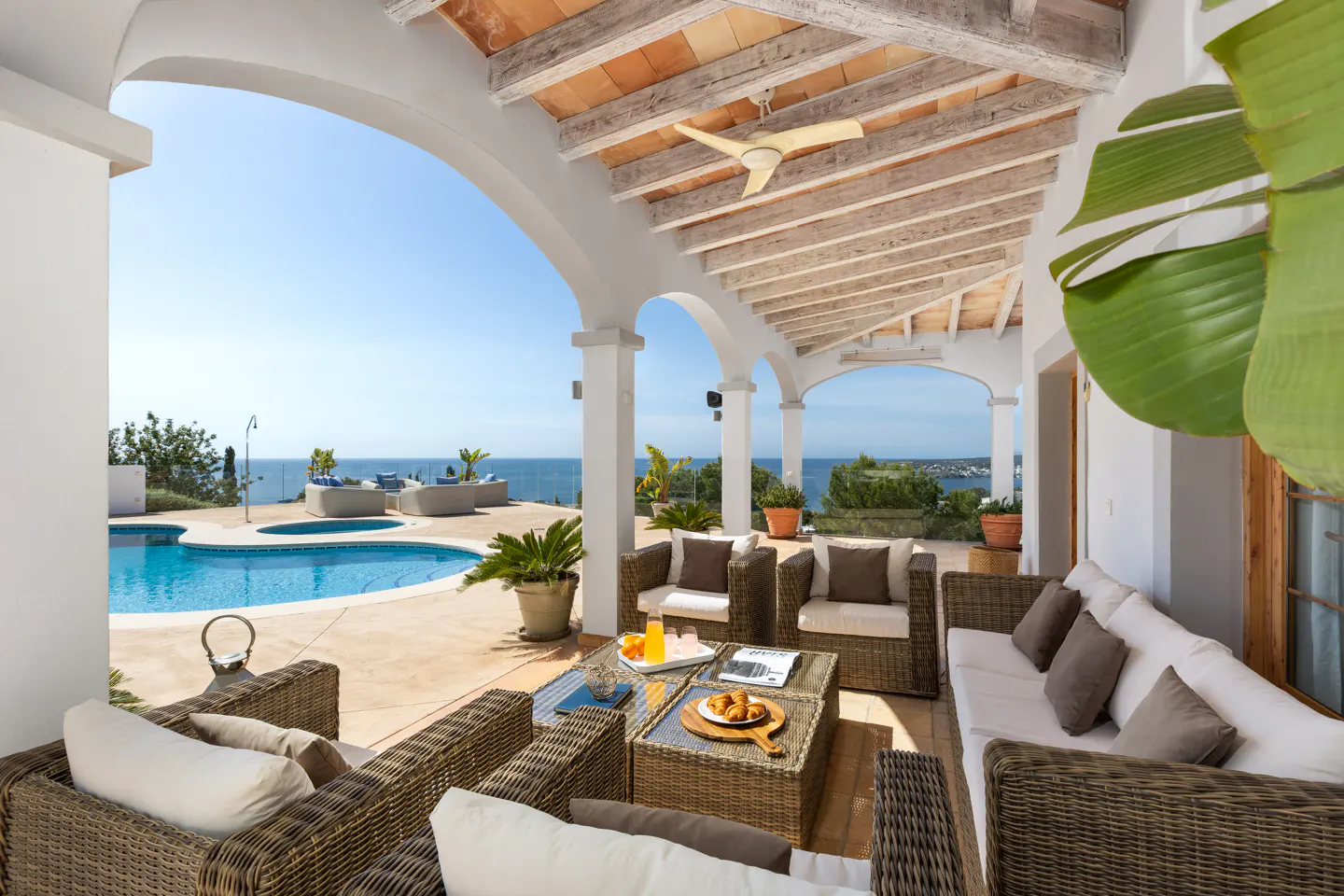 Outdoor patio with wicker furniture, white cushions, and a table with food and drinks. A pool and ocean are visible in the background.