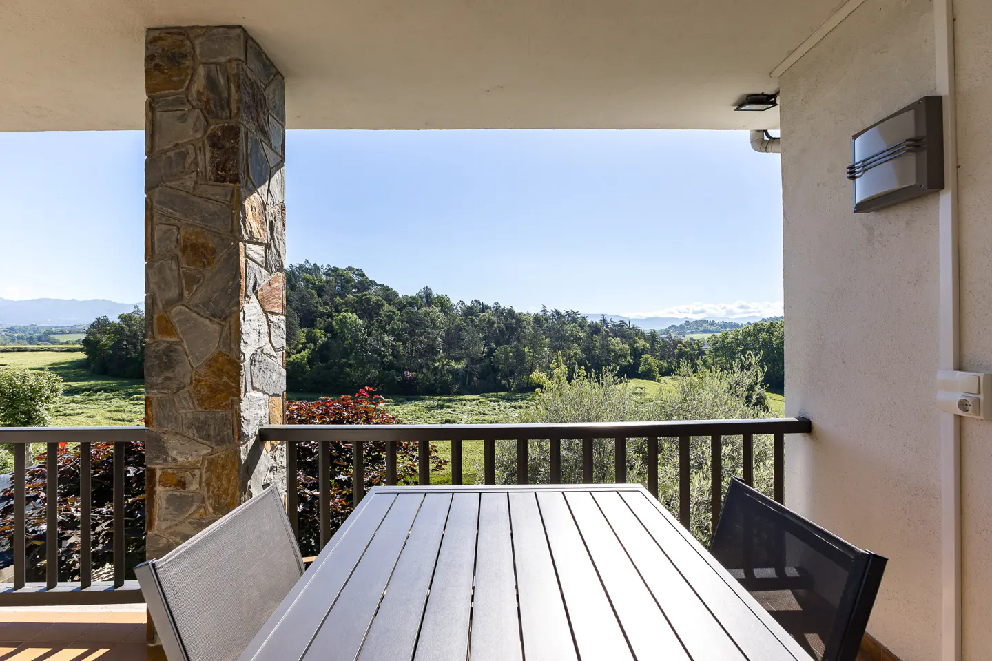 Balcony view with table, chairs, and stone pillar overlooking green fields and trees under a blue sky.