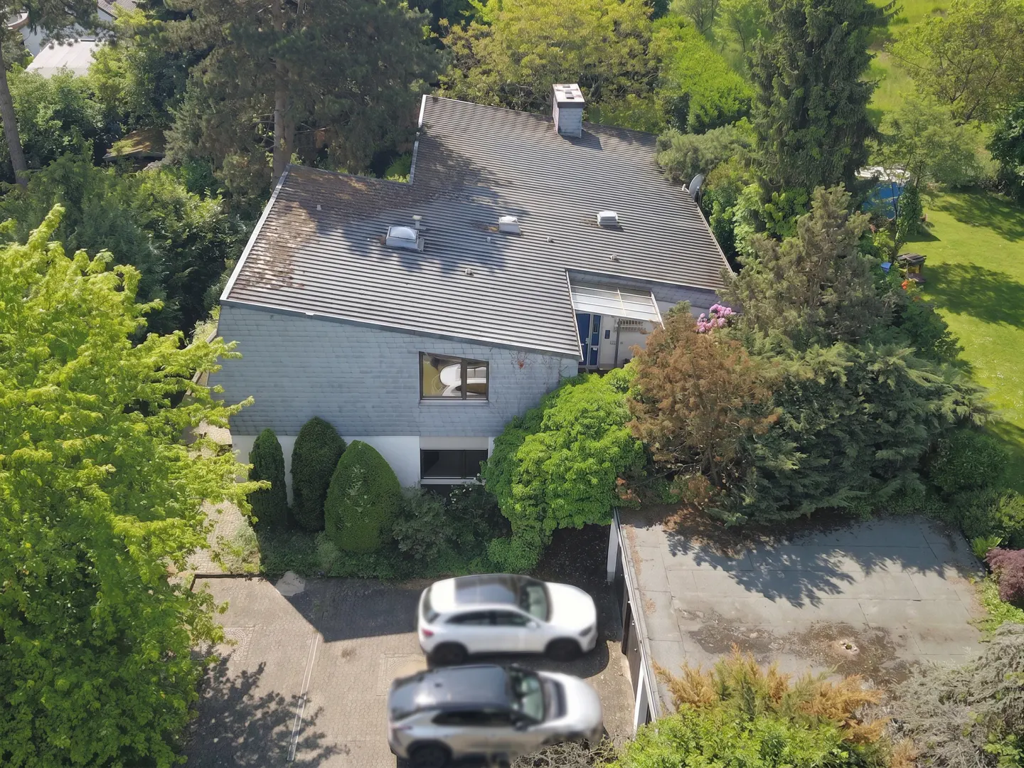Aerial view of a gray, modern house with a gray roof, surrounded by green trees and two parked cars.