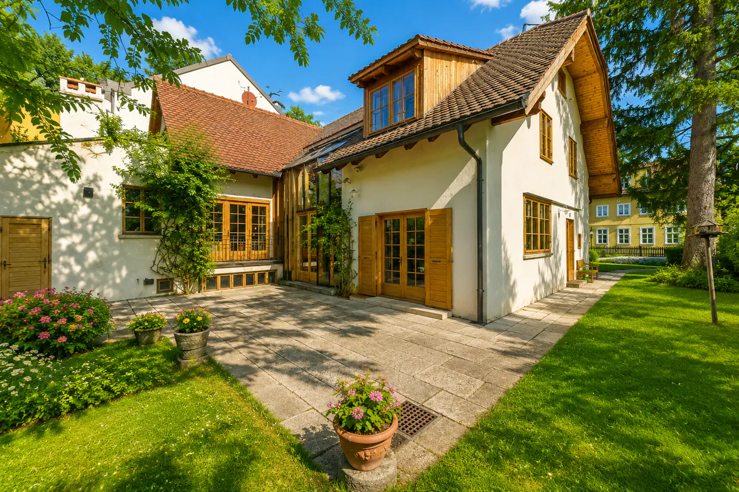 Exterior view of a cream-colored house with a brown roof, wooden windows, and a stone patio surrounded by green grass and trees.