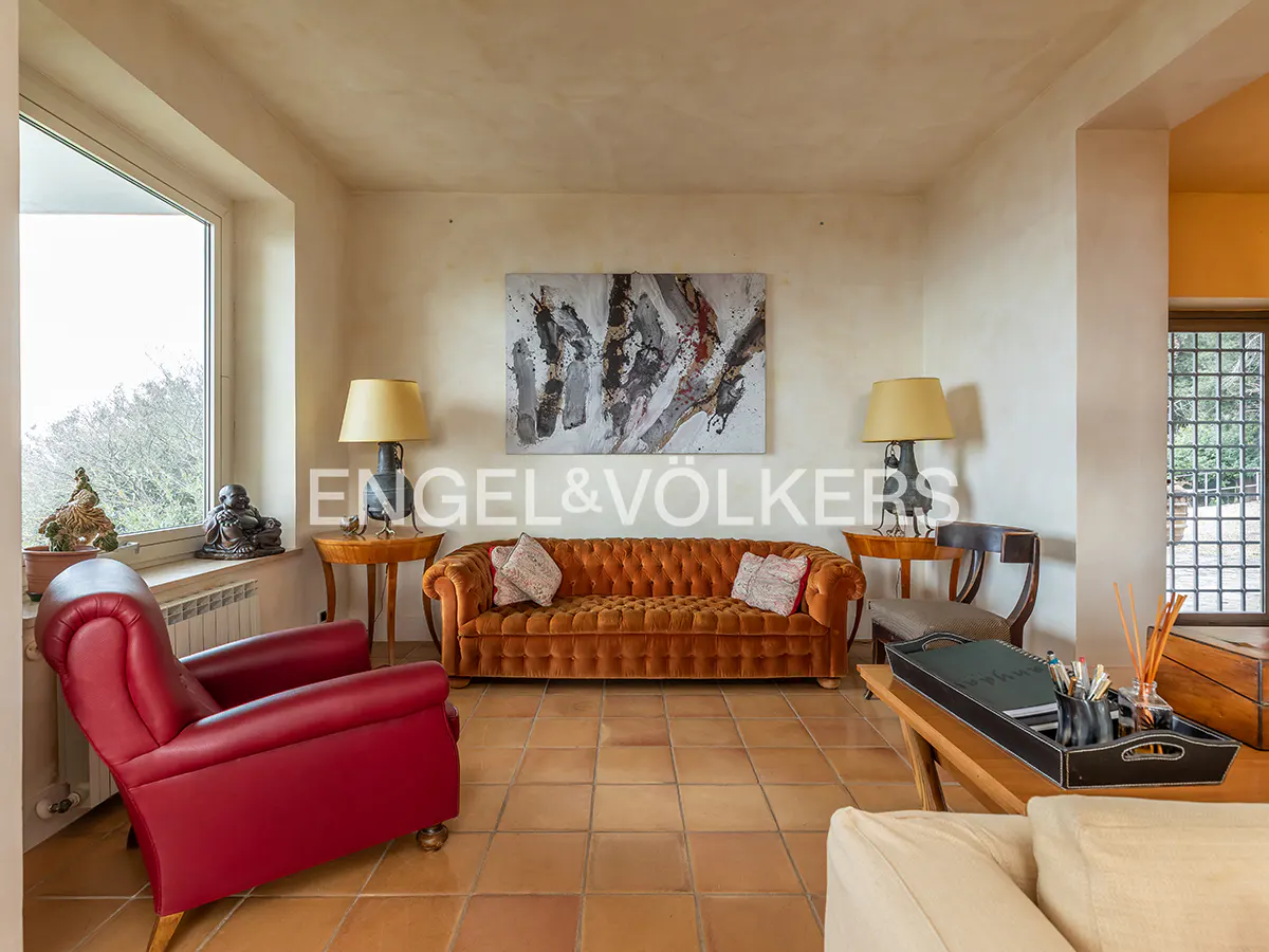 Living room with terracotta tile floor, orange sofa, red chair, and abstract art above the sofa. Engel & Völkers logo visible.
