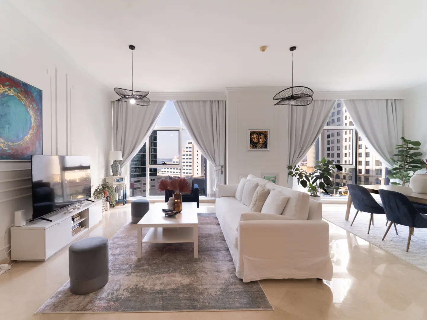 Bright living room with white sofa, coffee table, TV, and city view through large windows with gray curtains. Modern pendant lights hang above.