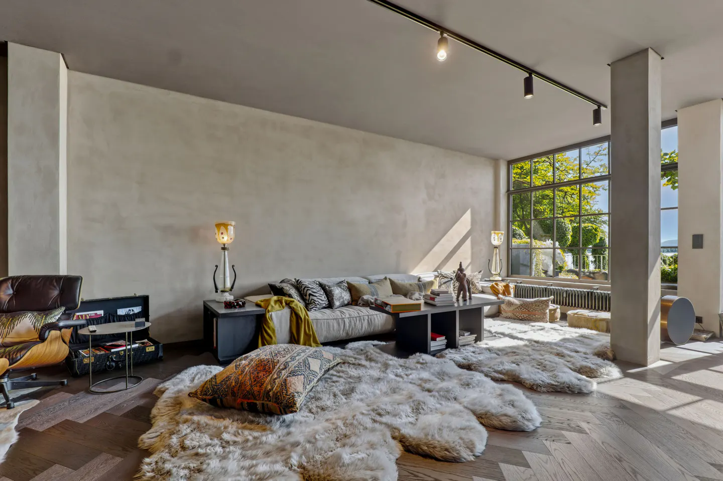 Modern living room with a beige sofa, fur rugs on herringbone wood floors, track lighting, and large windows with greenery.