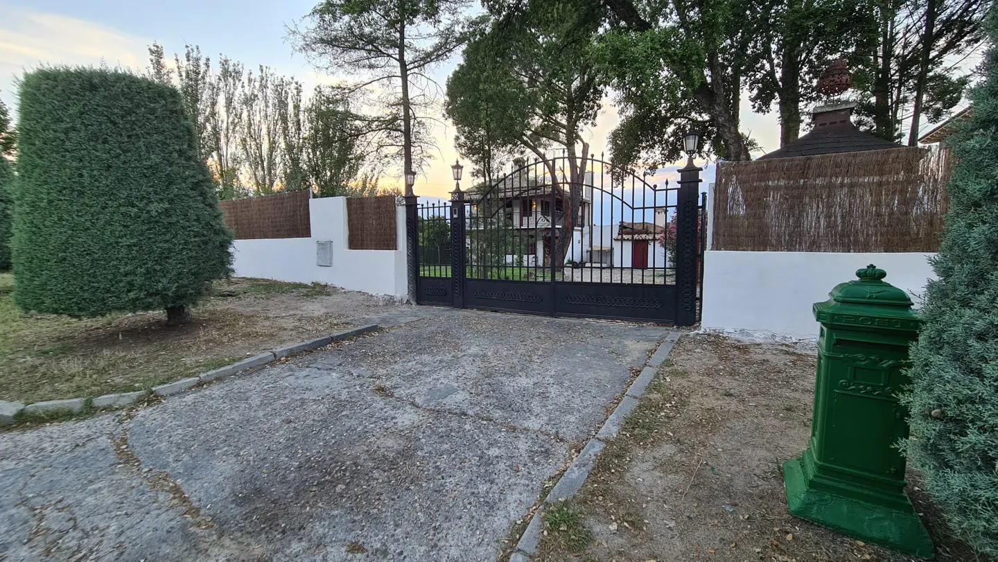 A paved driveway leads to a black metal gate with a white wall, green mailbox, and trees in the background.