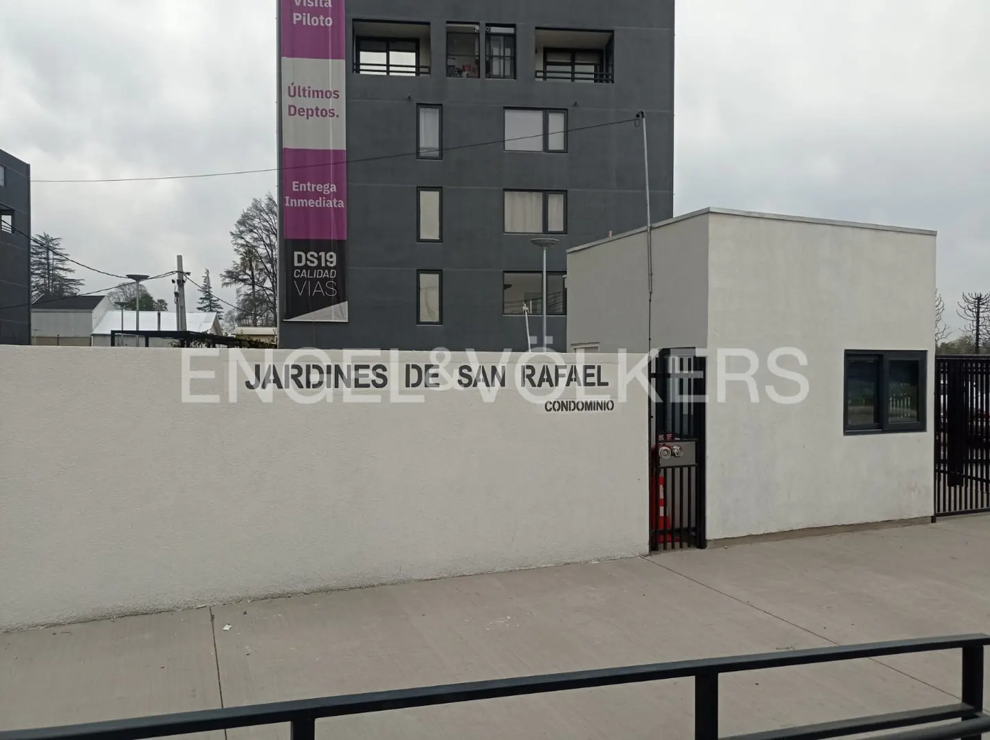White wall with "Jardines de San Rafael" sign in front of a modern gray apartment building under a cloudy sky.