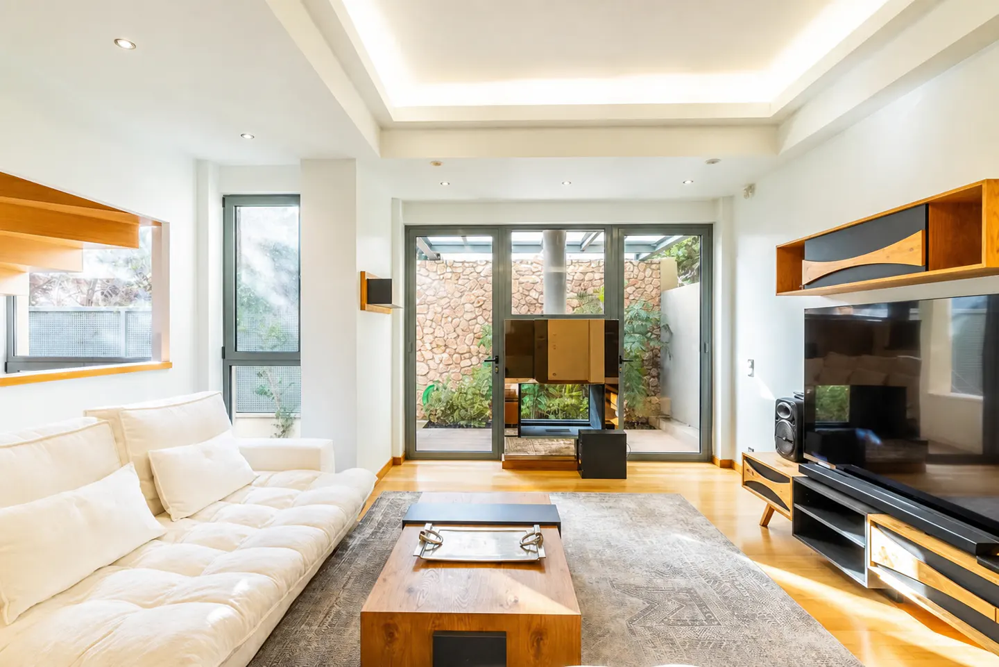 Modern living room with white sofa, wooden coffee table, large TV, and glass doors opening to a stone-walled patio.