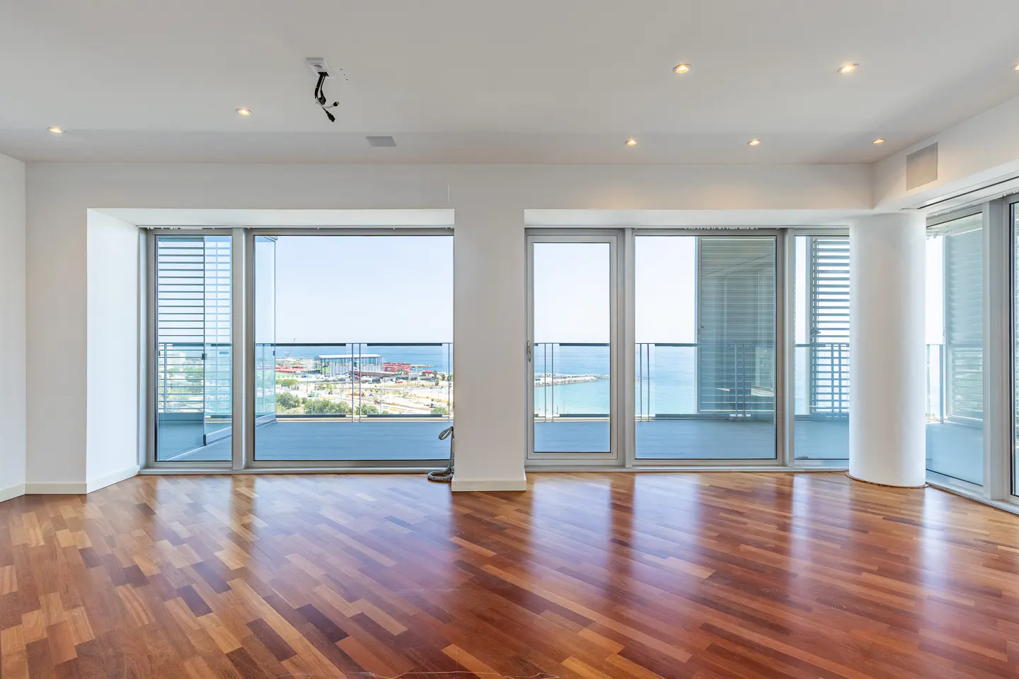 Empty room with polished hardwood floors, white walls, and large glass doors opening to a balcony with a coastal view.