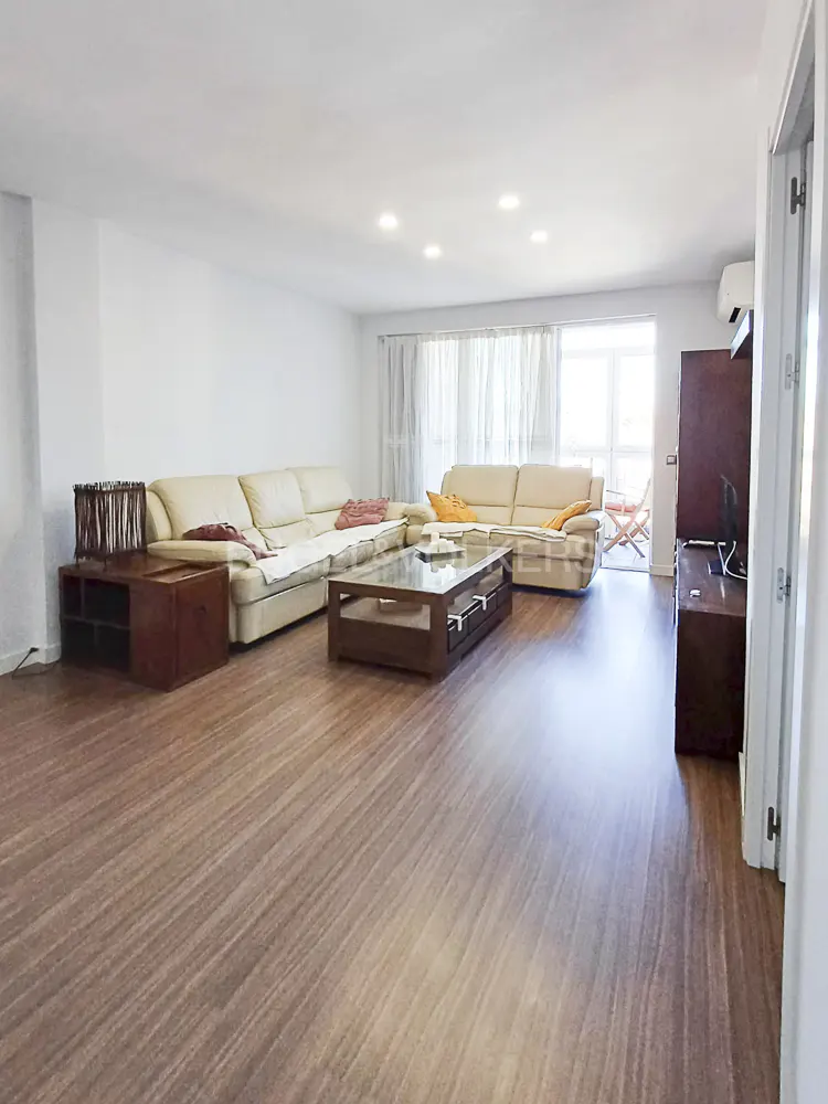 Bright living room with wood floors, white walls, and two cream-colored sofas facing each other. A glass-topped coffee table sits in the center.